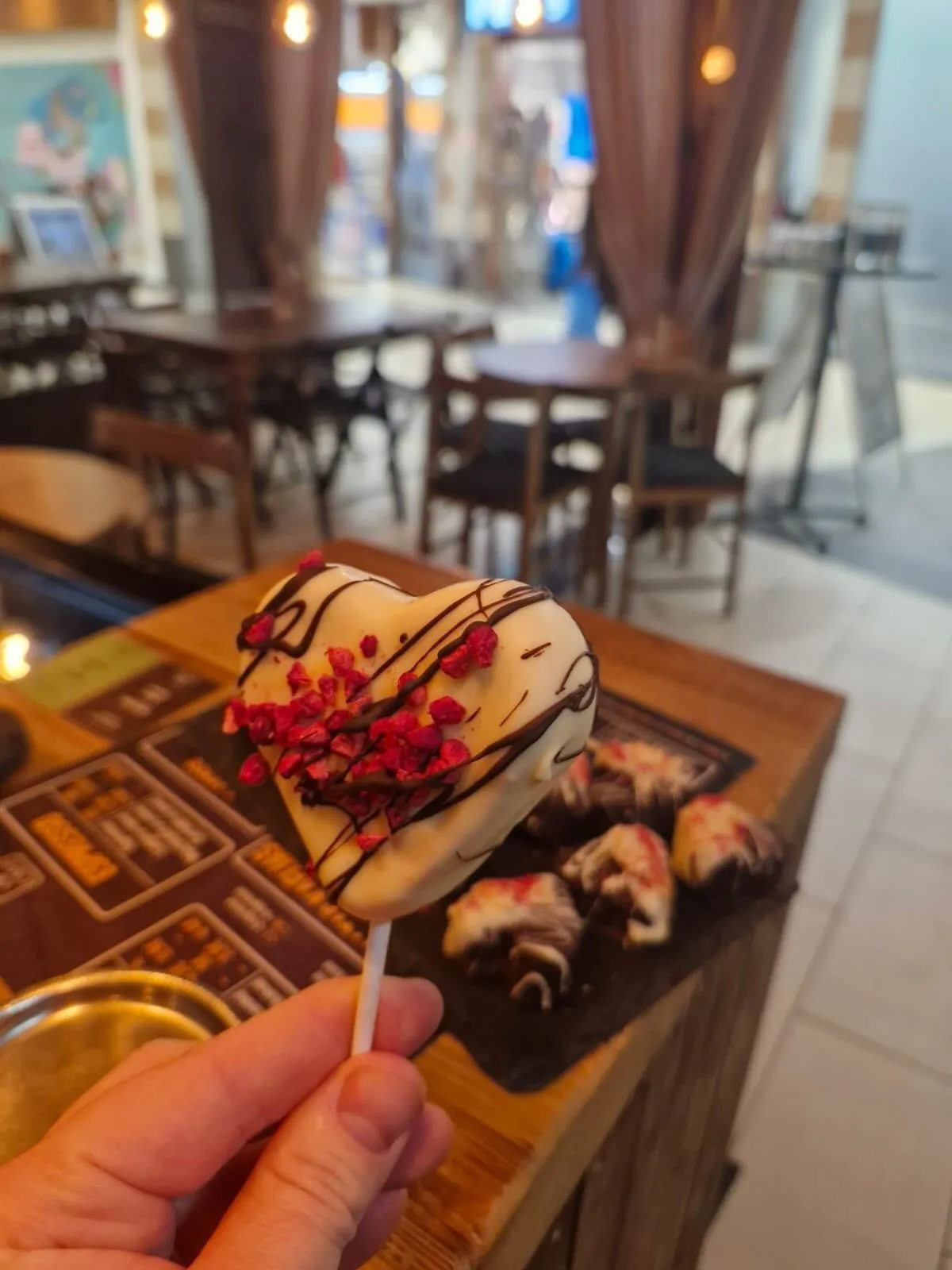 Sirsniņas formas kūciņa uz kociņa ar aveņu drupačām, fonā Kind of Cafe galdiņi